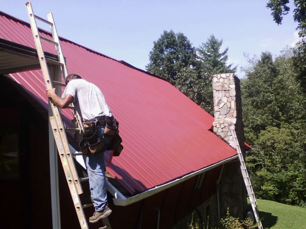 Roofer on a ladder beside a red metal roof and stone chimney — Soffit & Fascia Repair by Roofing Brothers in Kelley